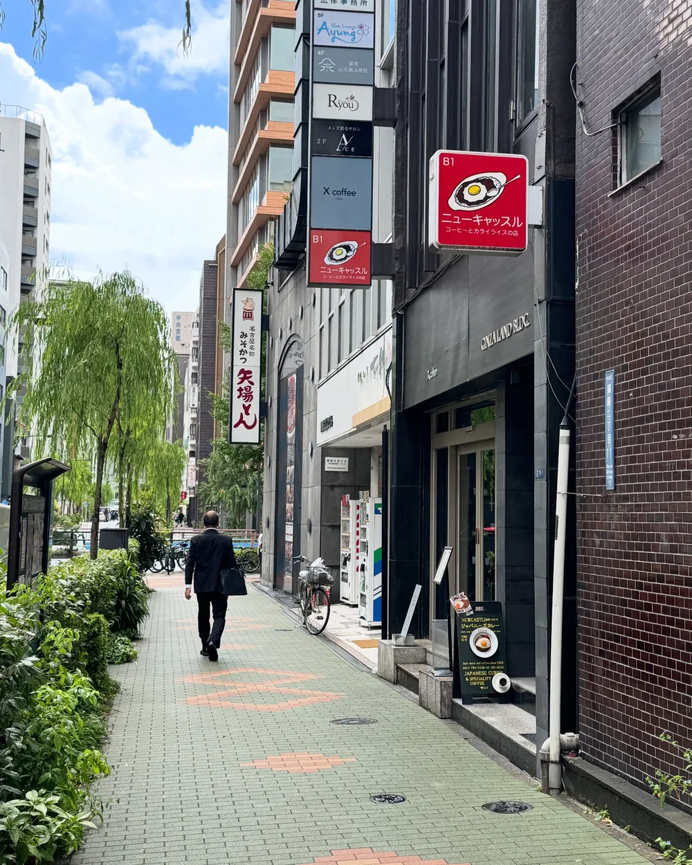 柳の並木がある歩道と、ビルの角に出たニューキャッスルの赤い突き出し看板